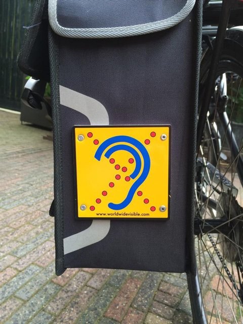 Limited Hearing reflecterend bordje gemonteerd op zijkant van fietstas met geel herkenningsteken Limited Hearing reflecterend bordje gemonteerd op zijkant van fietstas met geel herkenningsteken